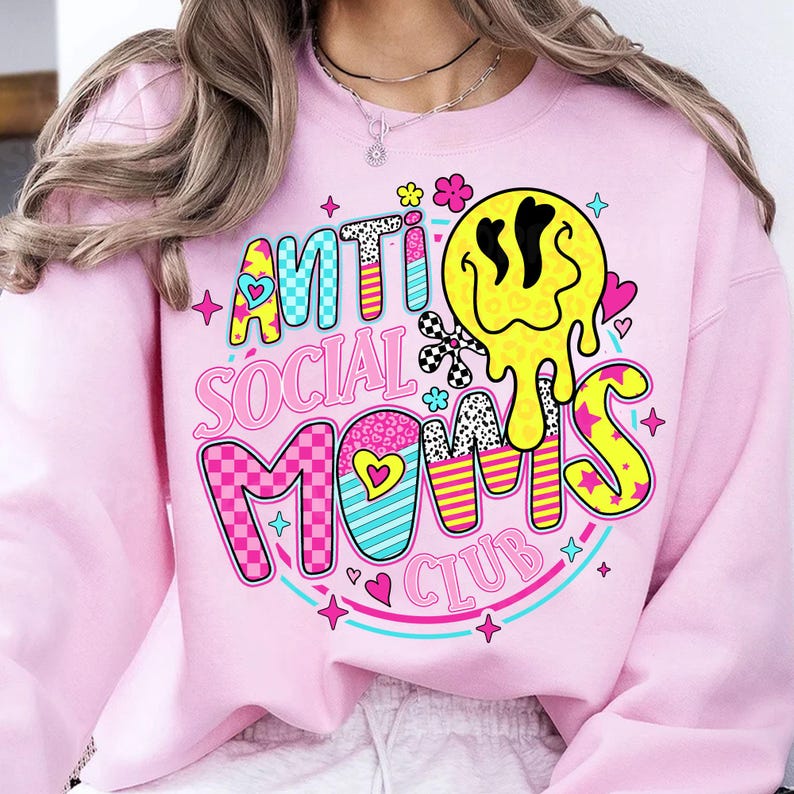 May include: A light pink sweatshirt with the text "ANTI SOCIAL MOMS CLUB" in a colorful, retro design. The graphic includes a melting yellow smiley face, hearts, stars, and checkered patterns. The sweatshirt is a casual, comfortable style.
