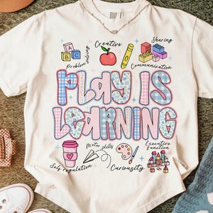 Puede incluir: Camiseta color crema con las palabras "PLAY IS LEARNING" en letras grandes y coloridas. El diseño incluye ilustraciones de bloques de construcción, una manzana y una taza de café, con palabras como "Creativo" y "Compartir".