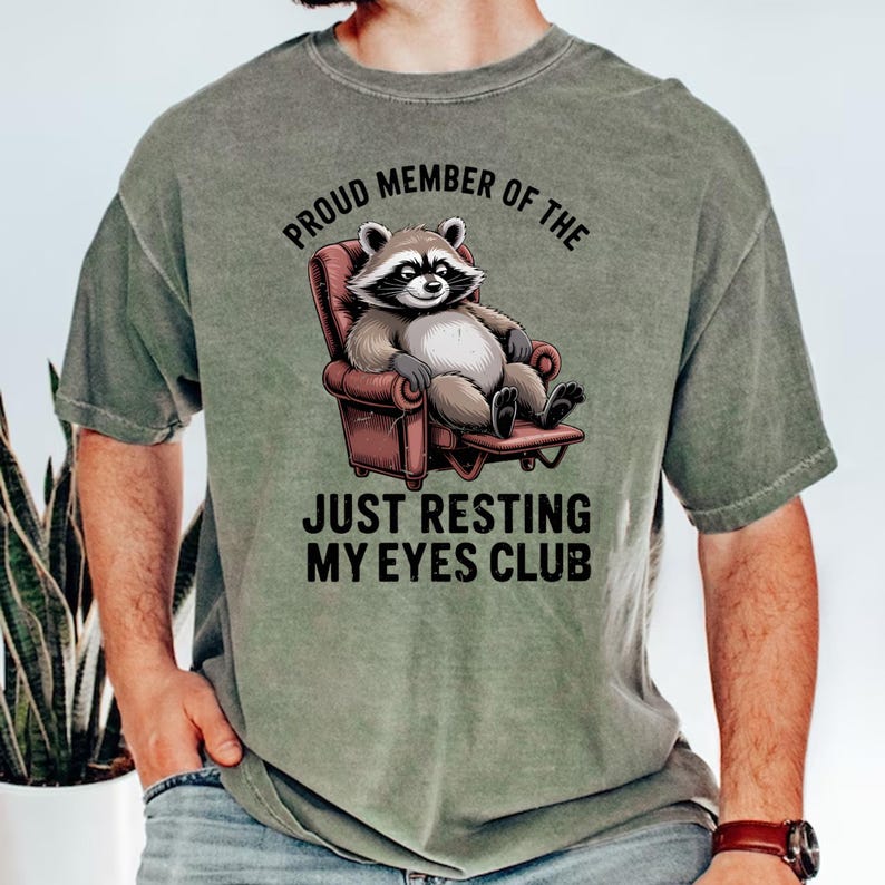 Op de afbeelding: Olijfgroen t-shirt met een cartoon wasbeer in een rode fauteuil. De tekst luidt "Proud Member of the Just Resting My Eyes Club". De wasbeer heeft een zwart masker en glimlacht.