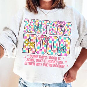 May include: A white sweatshirt with a checkered design that says "MOTHERHOOD" in pink, blue, and yellow letters. The text below the design says "SOME DAYS I ROCK IT. SOME DAYS IT ROCKS ME. EITHER WAY WE'RE ROCKIN'."