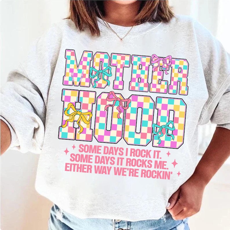 May include: A white sweatshirt with a checkered design that says "MOTHERHOOD" in pink, blue, and yellow letters. The text below the design says "SOME DAYS I ROCK IT. SOME DAYS IT ROCKS ME. EITHER WAY WE'RE ROCKIN'."