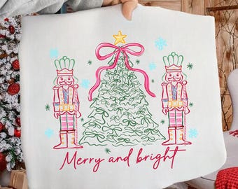 Può includere: Federa bianca a tema natalizio con un disegno disegnato a mano. Il disegno presenta un albero di Natale verde con un fiocco e una stella rosa, affiancato da due figure di schiaccianoci. Le parole "Merry and bright" sono scritte in rosso.