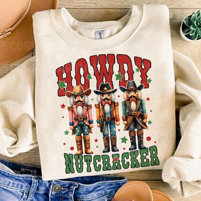 Puede incluir: Una sudadera blanca con un gr&aacute;fico rojo, verde y blanco de tres figuras de cascanueces vaquero. El texto "Howdy Nutcracker" est&aacute; impreso en verde y rojo.