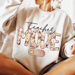 May include: White sweatshirt with the text "Teacher Mode Off" in pink and brown lettering with floral accents. The text "Off" is on a pink and brown toggle switch.