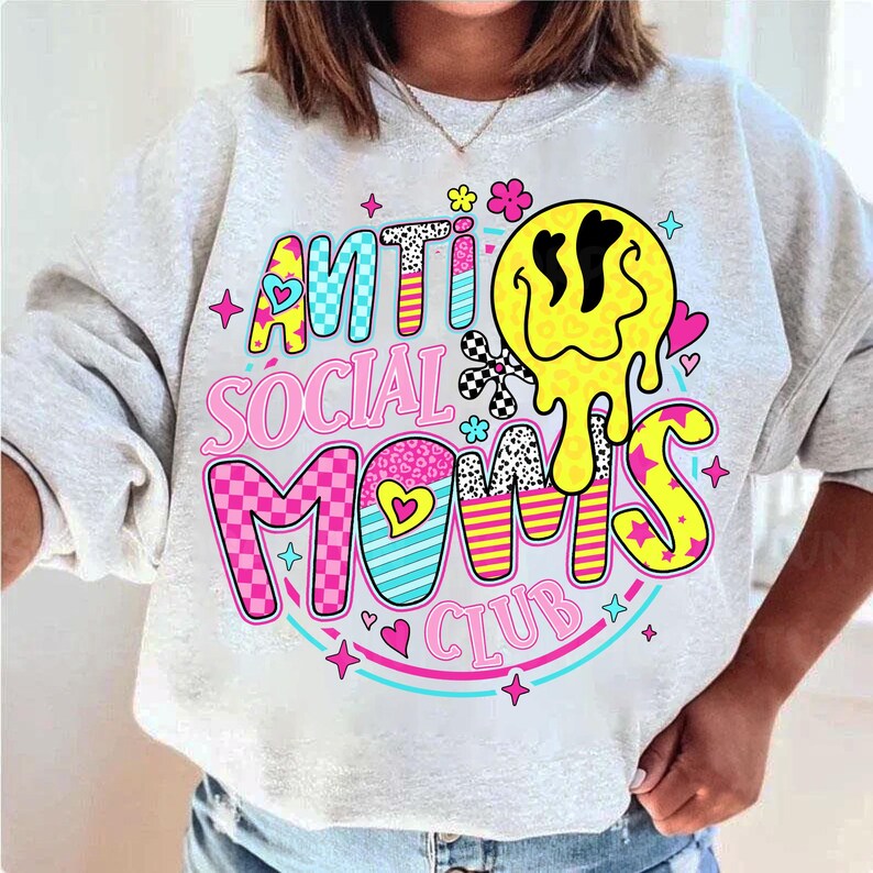 May include: A light gray sweatshirt with the text "ANTI SOCIAL MOMS CLUB" in a colorful, playful design. The design includes a yellow melting smiley face, hearts, stars, and floral accents. The sweatshirt is a casual, comfortable style.