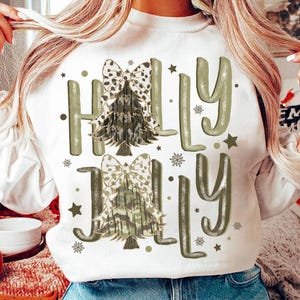 Può includere: Felpa bianca con un design di albero di Natale mimetico verde e bianco e il testo "Holly Jolly" in lettere glitter verdi.