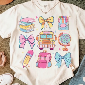 May include: Cream-colored t-shirt featuring colorful school-themed illustrations. Designs include books, bows, a school bus, a globe, a pencil, a backpack, and a box of crayons. The illustrations are outlined in black and filled with bright colors.