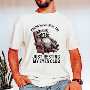 Op de afbeelding: Cr&egrave;mekleurig T-shirt met een cartoon wasbeer in een bruine fauteuil. De tekst luidt "PROUD MEMBER OF THE JUST RESTING MY EYES CLUB" in zwart, gebogen boven en onder de wasbeerillustratie.