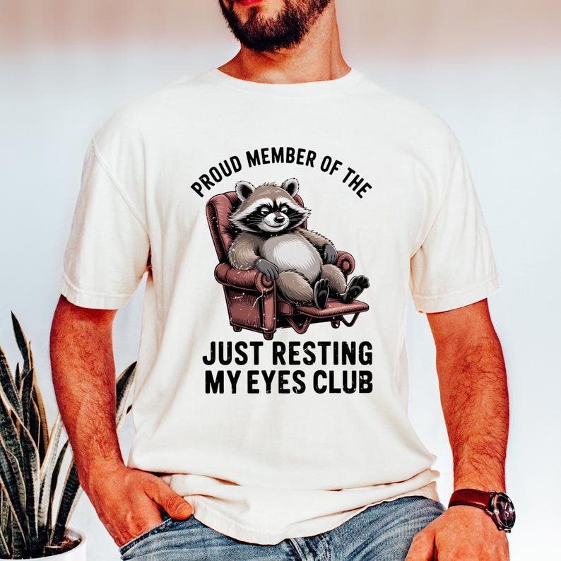 Op de afbeelding: Cr&egrave;mekleurig T-shirt met een cartoon wasbeer in een bruine fauteuil. De tekst luidt "PROUD MEMBER OF THE JUST RESTING MY EYES CLUB" in zwart, gebogen boven en onder de wasbeerillustratie.