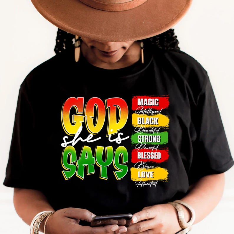 May include: Black t-shirt with the words "GOD she is SAYS" in yellow, red, and green. The shirt also features a list of words: Magic, Intelligent, Black, Beautiful, Strong, Powerful, Blessed, Brave, Love, and Influential.