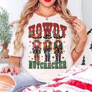 Puede incluir: Camiseta blanca con un gr&aacute;fico de tres cascanueces vaqueros y el texto "Howdy Nutcracker".