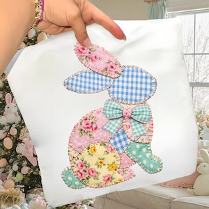 Op de afbeelding: Wit vierkant kussen met een patchwork konijn ontwerp. Het konijn is gemaakt van verschillende stoffen met patronen, waaronder roze bloemen, blauw gingham en mintgroene stippen. Een mintgroene strik siert de nek van het konijn en creëert een Paas thema.