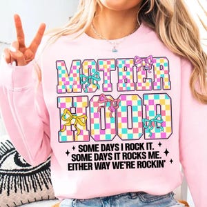 May include: Pink sweatshirt with a checkered design that says "MOTHERHOOD" in large letters with colourful bows. The text below says "SOME DAYS I ROCK IT. SOME DAYS IT ROCKS ME. EITHER WAY WE'RE ROCKIN'."
