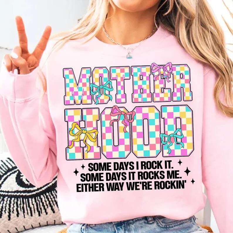 May include: Pink sweatshirt with a checkered design that says "MOTHERHOOD" in large letters with colourful bows. The text below says "SOME DAYS I ROCK IT. SOME DAYS IT ROCKS ME. EITHER WAY WE'RE ROCKIN'."