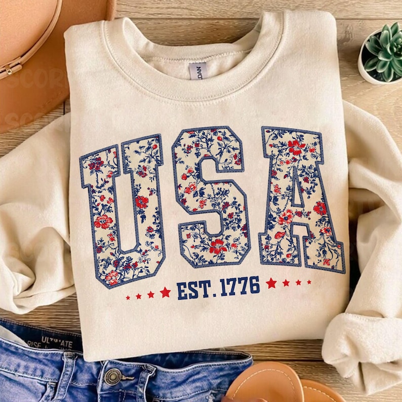 May include: A cream-colored sweatshirt with a floral print of the word "USA" in navy blue and red. The text "EST. 1776" is printed below the word "USA" in navy blue.