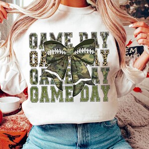 May include: White sweatshirt with the text "GAME DAY" repeated four times in green glitter letters. A green camouflage bow with white stripes and football laces is centered on the shirt.