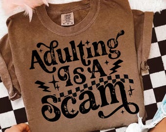 Könnte beinhalten: Braunes Comfort Colors T-Shirt mit dem schwarzen Schriftzug "Adulting is a Scam". Das Design umfasst Sterne, Blitze und ein Schachbrettmuster. Das T-Shirt ist gefaltet.