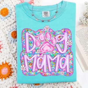 May include: A turquoise blue t-shirt with a pink paw print and the text "Dog Mama" surrounded by a floral design in shades of pink, purple, and green.