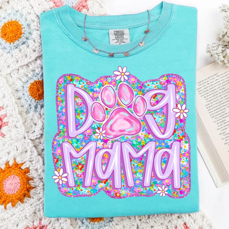 May include: A turquoise blue t-shirt with a pink paw print and the text "Dog Mama" surrounded by a floral design in shades of pink, purple, and green.