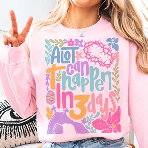 May include: Pink sweatshirt with a colorful graphic design that says "A lot can happen in 3 days". The graphic includes flowers, an Easter egg, a cross, and a tomb.