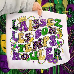 以下が含まれることがあります： 「Laissez les bons temps rouler」のフレーズが金、紫、緑のグリッター文字で描かれた白いトートバッグ。王冠、ビーズ、マスクのデザインが施され、すべてお祝いの色で、マルディグラに最適です。