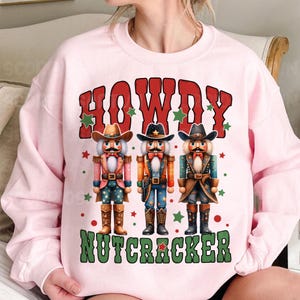 Puede incluir: Sudadera rosa con un dise&ntilde;o festivo con tres figuras de cascanueces vaquero y el texto "Howdy Nutcracker".