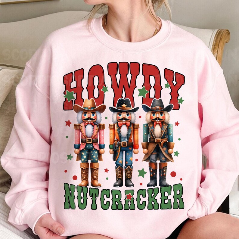 Puede incluir: Sudadera rosa con un dise&ntilde;o festivo con tres figuras de cascanueces vaquero y el texto "Howdy Nutcracker".
