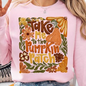 Pode incluir: Camisola rosa claro com a frase bordada "Take Me to the Pumpkin Patch". O design apresenta uma abóbora, flores e folhas em tons de laranja, castanho e verde. Uma peça de vestuário com tema de outono.