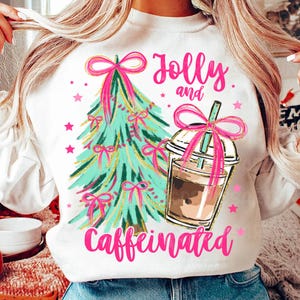 Puede incluir: Sudadera blanca con un árbol de Navidad rosa y verde y una taza de café rosa y marrón. El texto "Jolly and Caffeinated" está impreso en rosa.