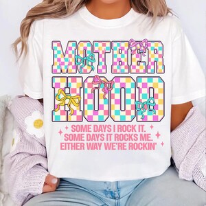 May include: White t-shirt with a checkered design that says "MOTHERHOOD" in pink, blue, and yellow. The text below says "SOME DAYS I ROCK IT. SOME DAYS IT ROCKS ME. EITHER WAY WE'RE ROCKIN'."
