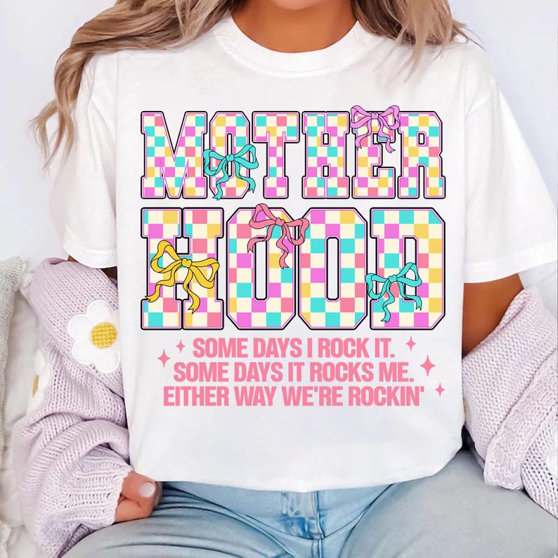 May include: White t-shirt with a checkered design that says "MOTHERHOOD" in pink, blue, and yellow. The text below says "SOME DAYS I ROCK IT. SOME DAYS IT ROCKS ME. EITHER WAY WE'RE ROCKIN'."