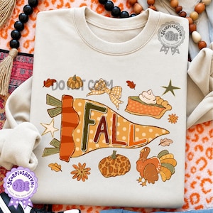 Pode incluir: Uma camisola bege de gola redonda com uma impressão gráfica de uma flâmula com a palavra FALL em letras grandes. O design inclui uma abóbora, uma fatia de tarte, um peru, um laço xadrez, estrelas e folhas de outono em tons de laranja e castanho.