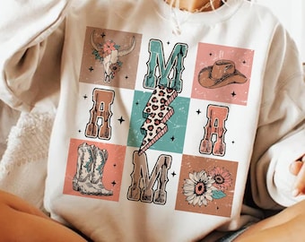 Puede incluir: Una sudadera blanca con un diseño gráfico de la palabra "MAMA" en estilo vintage. Las letras están rodeadas de varias imágenes, incluyendo una calavera, botas de vaquero, un sombrero de vaquero y girasoles.