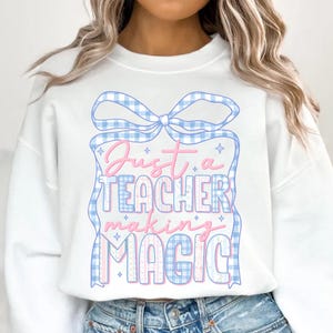 Puede incluir: Sudadera blanca con un gráfico azul y rosa. El gráfico dice "Just a TEACHER making MAGIC" en una fuente juguetona, rodeado por un lazo y marco de cuadros azules. Una parte superior cómoda y elegante para profesores.