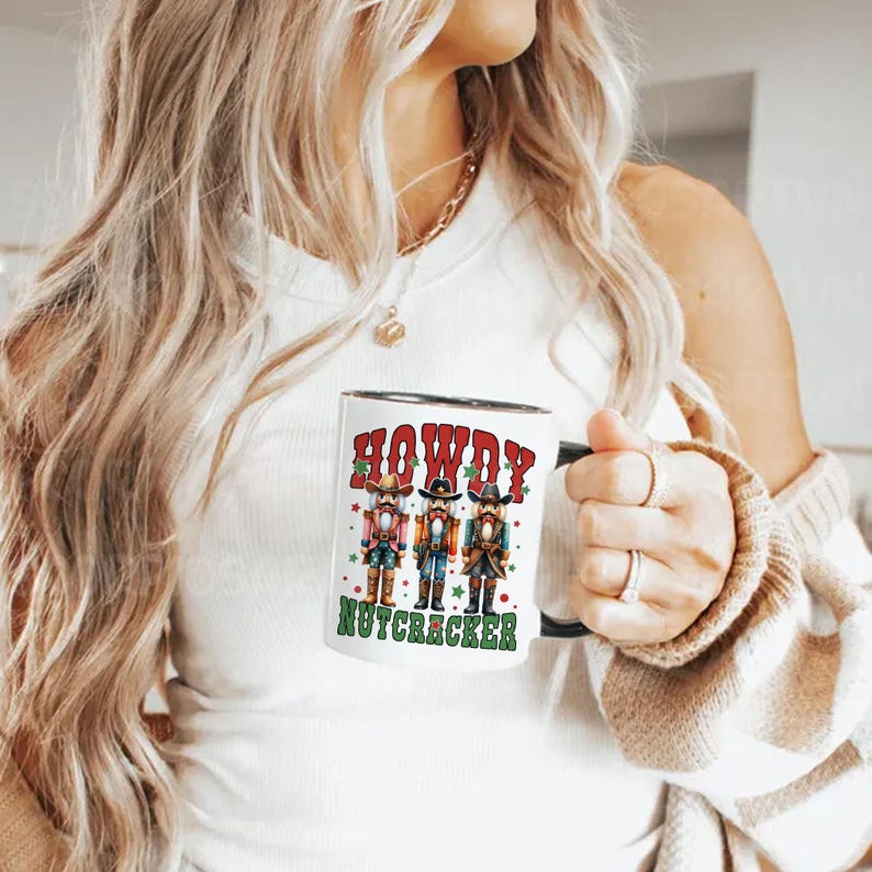 Puede incluir: Taza de cer&aacute;mica blanca con borde y asa negros. La taza presenta un gr&aacute;fico de tres cascanueces vaqueros con el texto "Howdy Nutcracker" en rojo y verde.