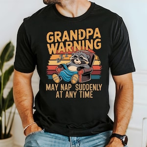 Könnte beinhalten: Schwarzes T-Shirt mit einer Grafik, die einen Waschbären in einem Sessel, einer Decke und dem Text "GRANDPA WARNING MAY NAP SUDDENLY AT ANY TIME" in Orange und Weiß zeigt. Das Design beinhaltet einen Sonnenuntergangshintergrund.