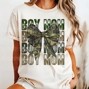 May include: A white t-shirt with the text "Boy Mom" repeated three times in a distressed font. A large camouflage bow is centered on the shirt.