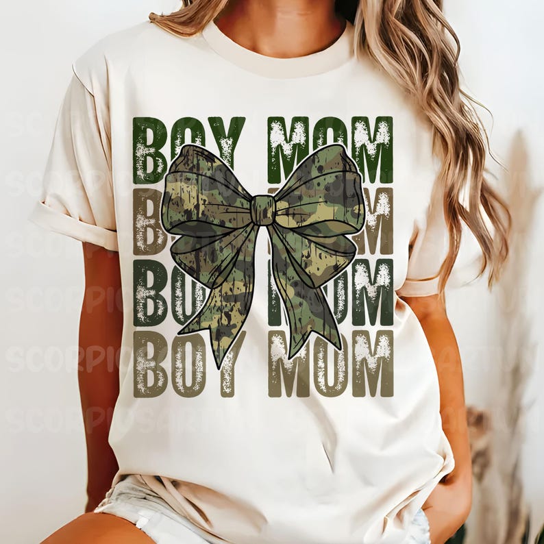 May include: A white t-shirt with the text "Boy Mom" repeated three times in a distressed font. A large camouflage bow is centered on the shirt.