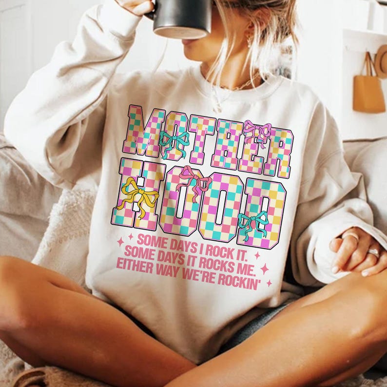 May include: A white sweatshirt with a checkered design and the text "MOTHERHOOD" in pink, blue and yellow. The text is surrounded by pink and yellow bows. The text below the design reads "SOME DAYS I ROCK IT. SOME DAYS IT ROCKS ME. EITHER WAY WE'RE ROCKIN'."