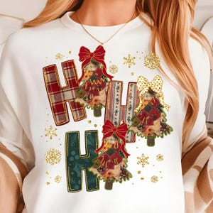 May include: White long-sleeve shirt with the text "HA HA HA" in a plaid and patterned design. The letters feature Christmas tree designs with red bows and gold snowflakes.