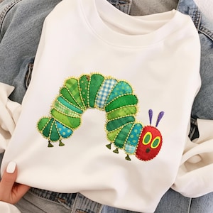 Puede incluir: Sudadera blanca con un diseño de oruga aplicado. La oruga está hecha de telas verdes, azules y amarillas, con una cabeza roja y ojos amarillos. El diseño está cosido con hilo dorado.