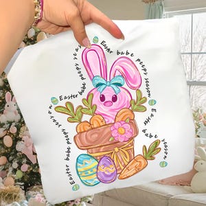 Może przedstawiać: Biała kwadratowa poduszka z różowym wzorem zajączka wielkanocnego. Zajączek znajduje się w brązowej doniczce z marchewkami i kwiatkiem. Poduszka ma napis "Easter babe peeps season" wokół zajączka i jajek.