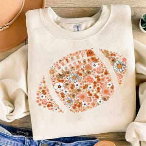 Puede incluir: Sudadera crema con un diseño floral de fútbol americano. El diseño está compuesto por flores naranjas, blancas y marrones. La sudadera tiene cuello redondo y es una prenda casual y cómoda.