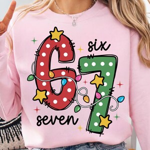 Könnte beinhalten: Hellrosa Sweatshirt mit den Zahlen "67" in Rot und Grün, verziert mit weißen Punkten, Sternen und Lichterketten. Das Wort "six" steht über der "6" und "seven" unter der "7".