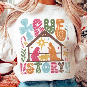 May include: White sweatshirt with a colorful graphic design featuring the words "TRUE STORY" and a stylized nativity scene. The design includes a star, candy canes, and other festive elements.