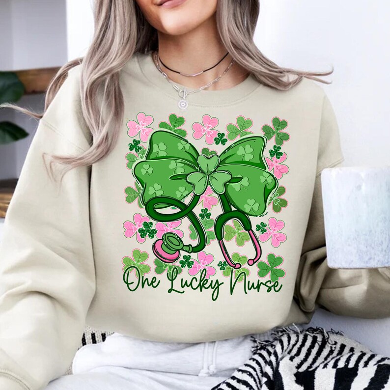 May include: A beige sweatshirt featuring a green bow and stethoscope design with pink accents and shamrocks. The text "One Lucky Nurse" is written in green script below the design. The sweatshirt is a casual style.