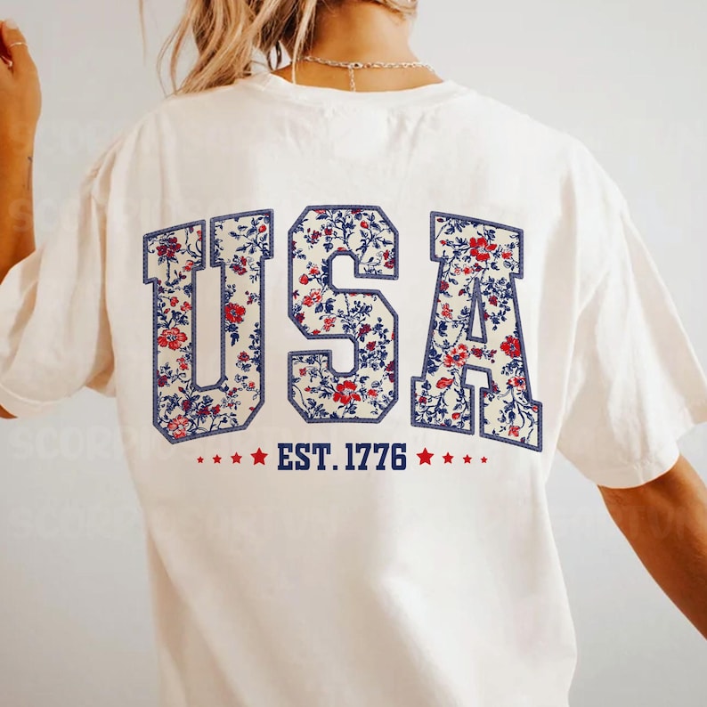 May include: White t-shirt with a floral print "USA" graphic on the back. The graphic also includes the text "EST. 1776" with red stars.