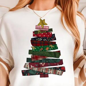 May include: White sweatshirt featuring a Christmas tree design. The tree is constructed from layered fabric strips in various patterns, including plaid, polka dots, and floral prints. A gold star tops the tree, creating a festive holiday theme.