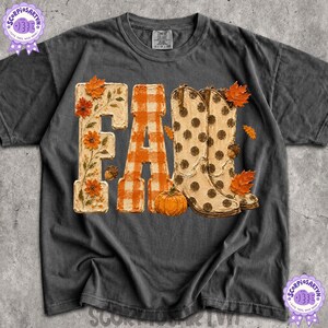 Puede incluir: Una camiseta de manga corta gris carbón con un estampado gráfico de la palabra FALL. La letra F está decorada con flores naranjas, la A es de cuadros naranjas y blancos, y las L son botas vaqueras beige con lunares marrones y una bellota.