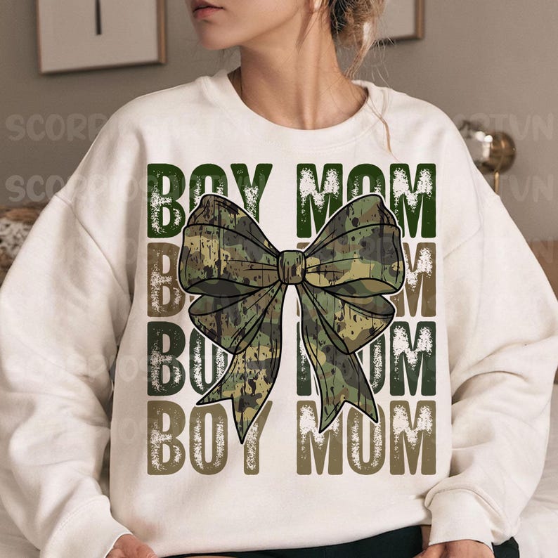 May include: White sweatshirt with the text "Boy Mom" repeated four times in a distressed font. A large camouflage bow is centered on the sweatshirt.
